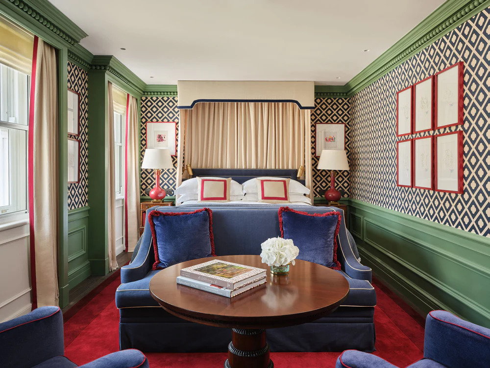 Traditional English hotel bedroom with green paneled walls, navy diamond-pattern wallpaper, a canopy bed, red-and-navy accent pillows, and a red patterned carpet. interior designers