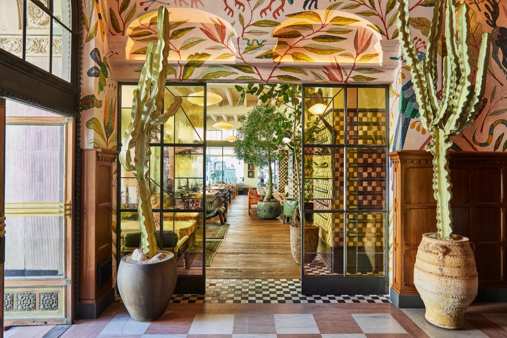 A sunlit hotel doorway framed by tropical murals and tall potted cacti, leading into a lush, plant-filled restaurant at LA Proper Hotel.