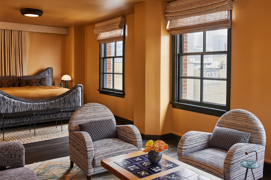 A spacious hotel suite featuring warm yellow walls, blue patterned armchairs, and a cozy king bed at LA Proper Hotel.