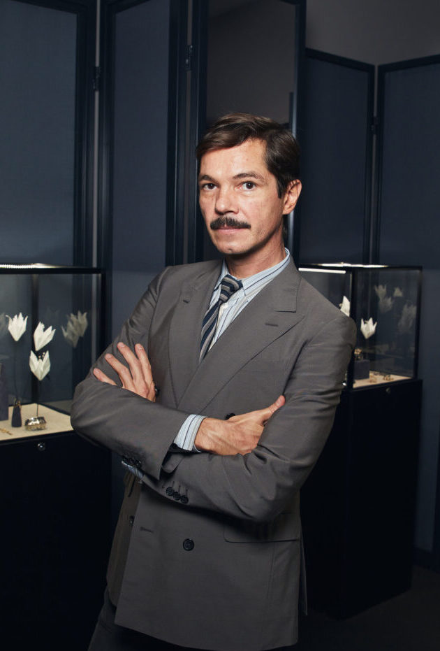 A portrait of a man with a mustache wearing a grey double-breasted suit, standing with crossed arms in front of jewelry display cases at PAD Saint-Tropez 2026