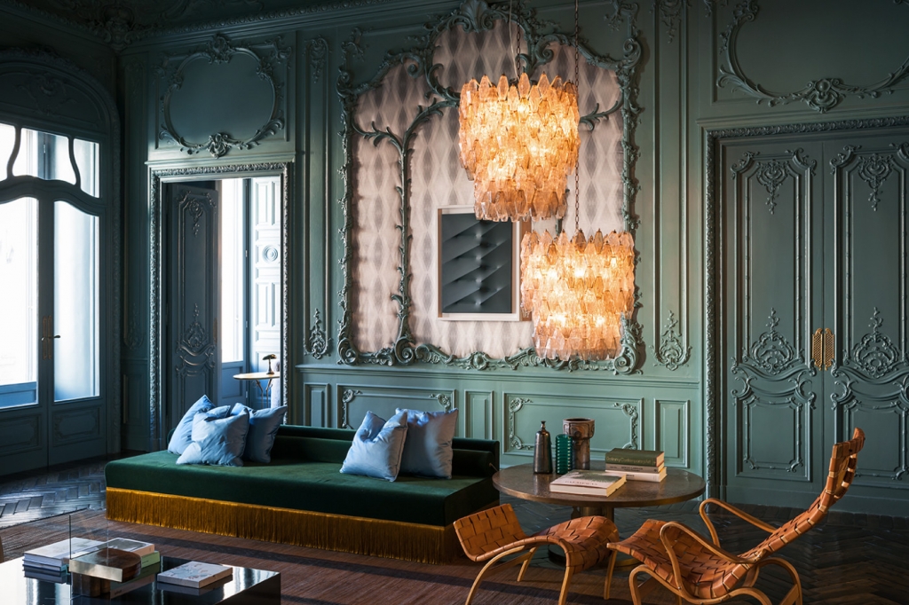 Ornate green boiserie-paneled salon with a fringed emerald velvet daybed, sky-blue cushions, woven leather lounge chairs, and an amber Murano glass chandelier set within an arched mirrored niche. interiors designers