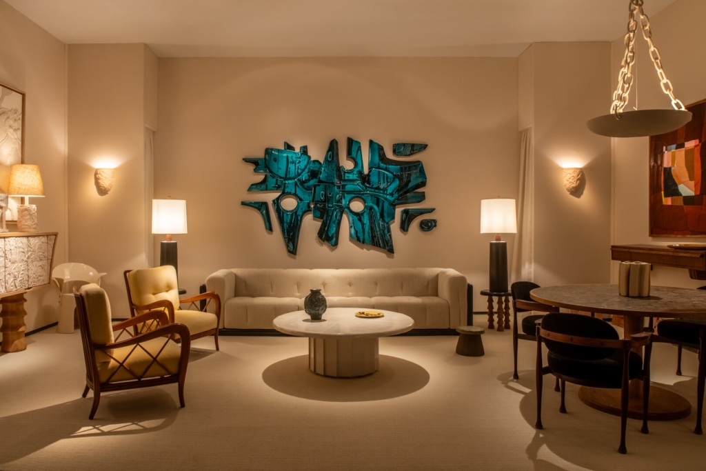 An elegant mid-century modern interior living room booth featuring a large teal abstract wall sculpture, cream furniture, and ambient lighting at PAD Saint-Tropez 2026.