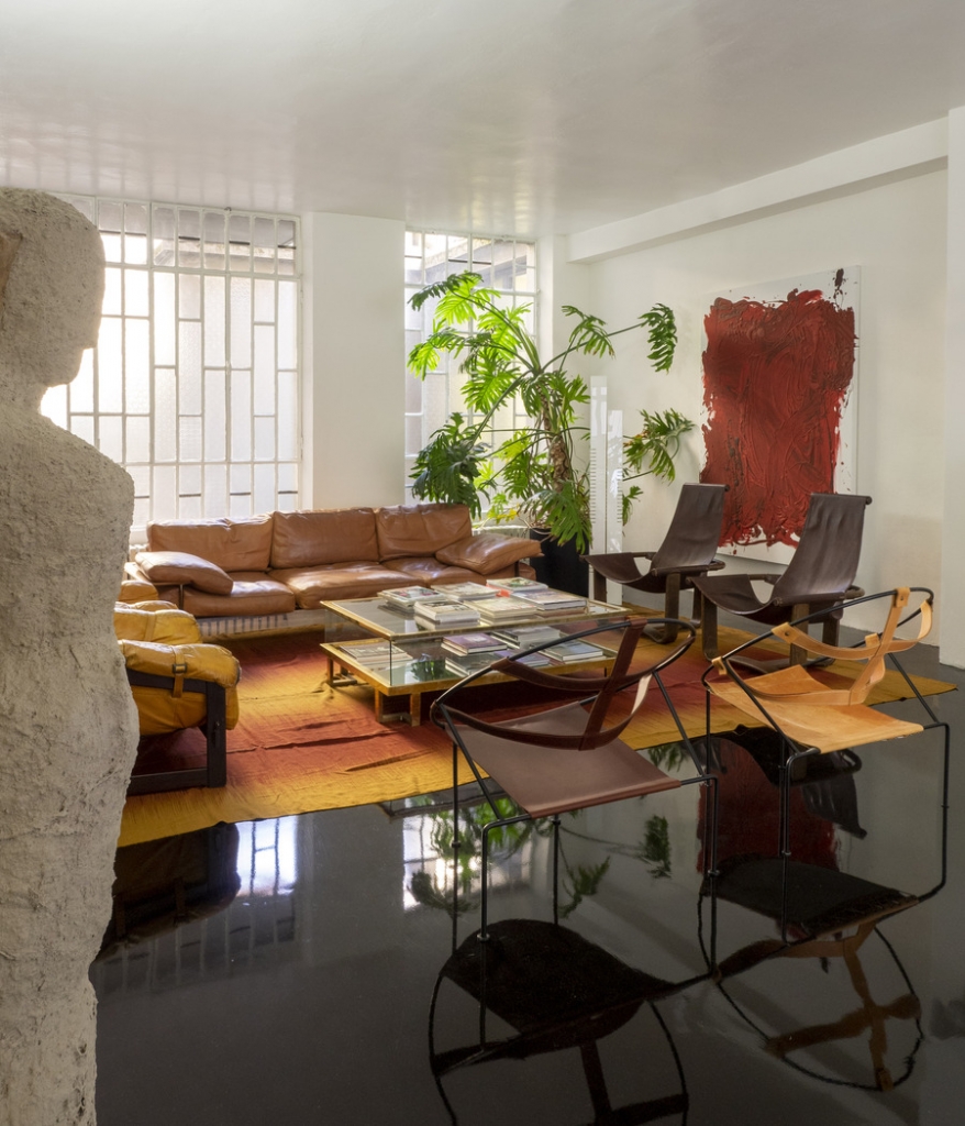 Sunlit loft-style living room with polished dark floors, camel leather sofas, sling-back leather and canvas chairs, a large red abstract painting, and a stone sculpture in the foreground. interior designers