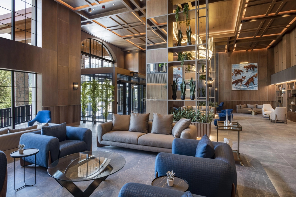 Warm wood-paneled hotel lobby with a mirrored plant divider, navy and taupe upholstered seating, a glass coffee table, and framed animal artwork on the wall. interior designers