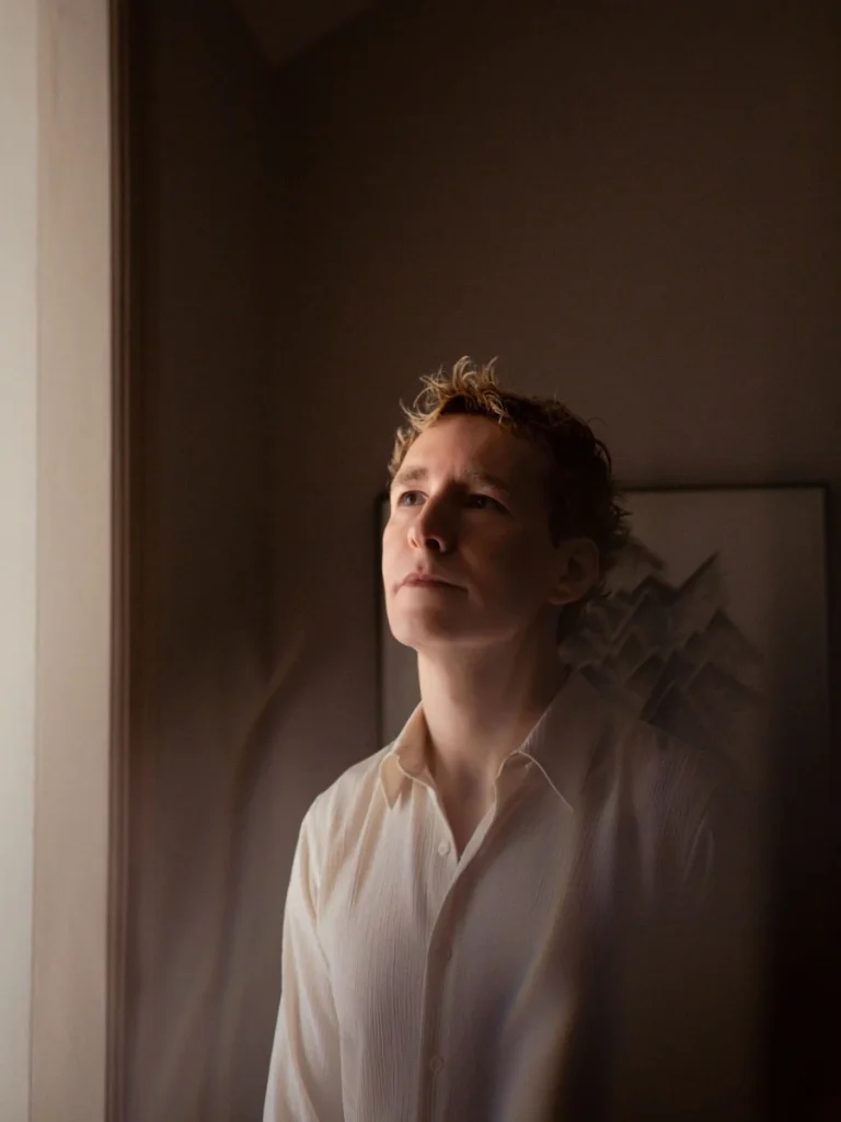 A portrait of a fair-haired person in a light-colored shirt looking upward near a window in softly lit room at PAD Saint-Tropez 2026