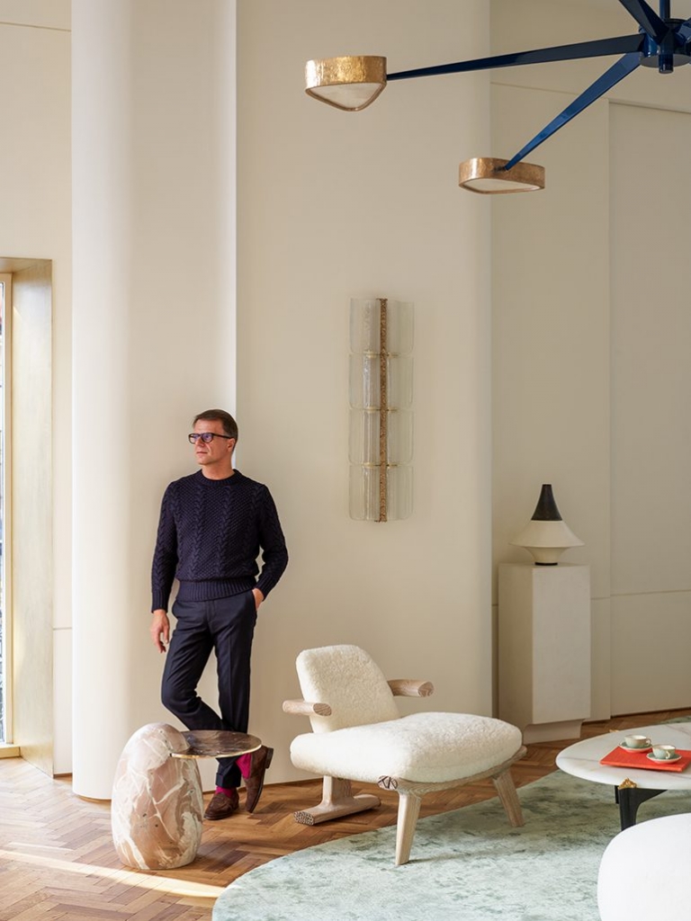 A man in a dark knit sweater leaning against a column in a brightly lit modern room with a cream armchair and unique light fixtures at PAD Saint-Tropez 2026.
