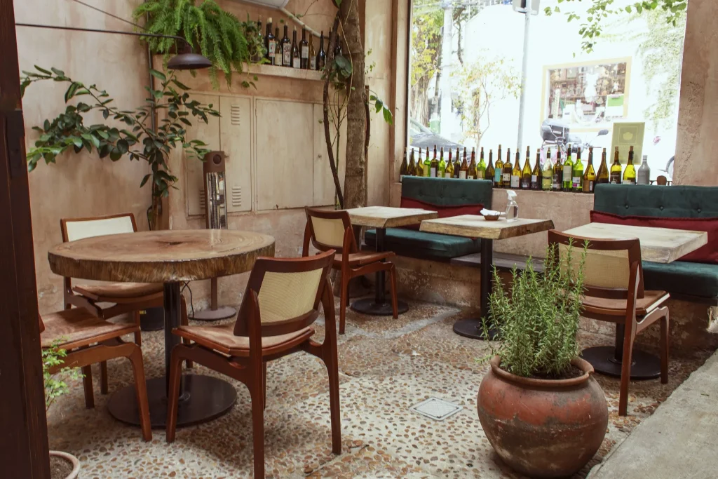 Área externa aconchegante com mesa de tronco de madeira, plantas, vaso de barro grande e prateleira com garrafas de vinho junto à janela - melhores restaurantes em São Paulo