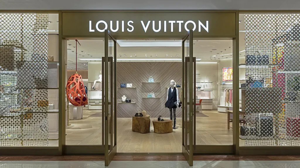 Entrada da boutique Louis Vuitton com portas abertas, piso de madeira clara, malas expostas e decoração em padrão monograma nas paredes - lojas de luxo em São Paulo
