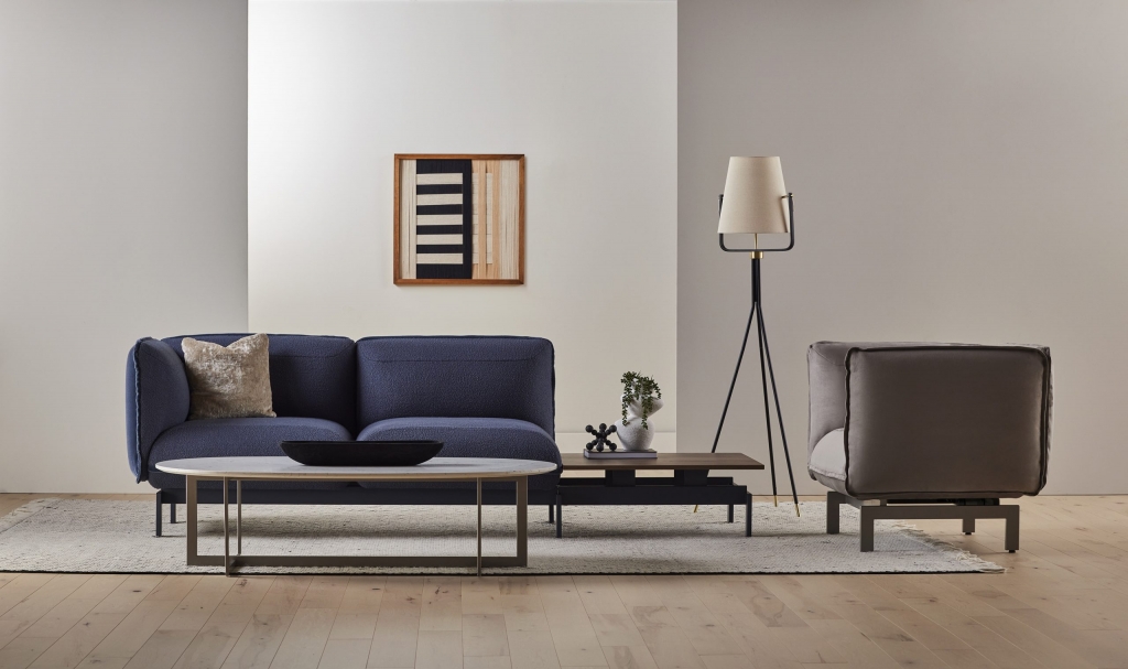 Minimalist furniture vignette featuring a navy boucle modular sofa, marble-topped coffee table, tripod floor lamp, and abstract woven wall art against a two-tone backdrop. interior designers