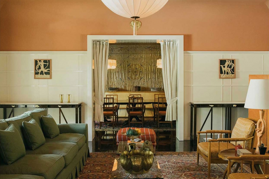 Warm bohemian bedroom nook with arched wicker-and-wood wall paneling, patterned drapery, marble coffee tables, and a tiled floor, opening to a balcony with tropical greenery. interior designers