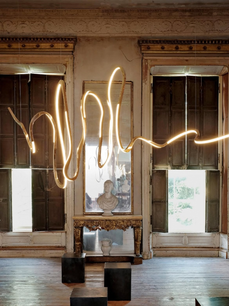 A dynamic, wave-like glowing LED and polished bronze light fixture suspended in front of old shuttered windows in a rustic room at PAD Saint-Tropez 2026.