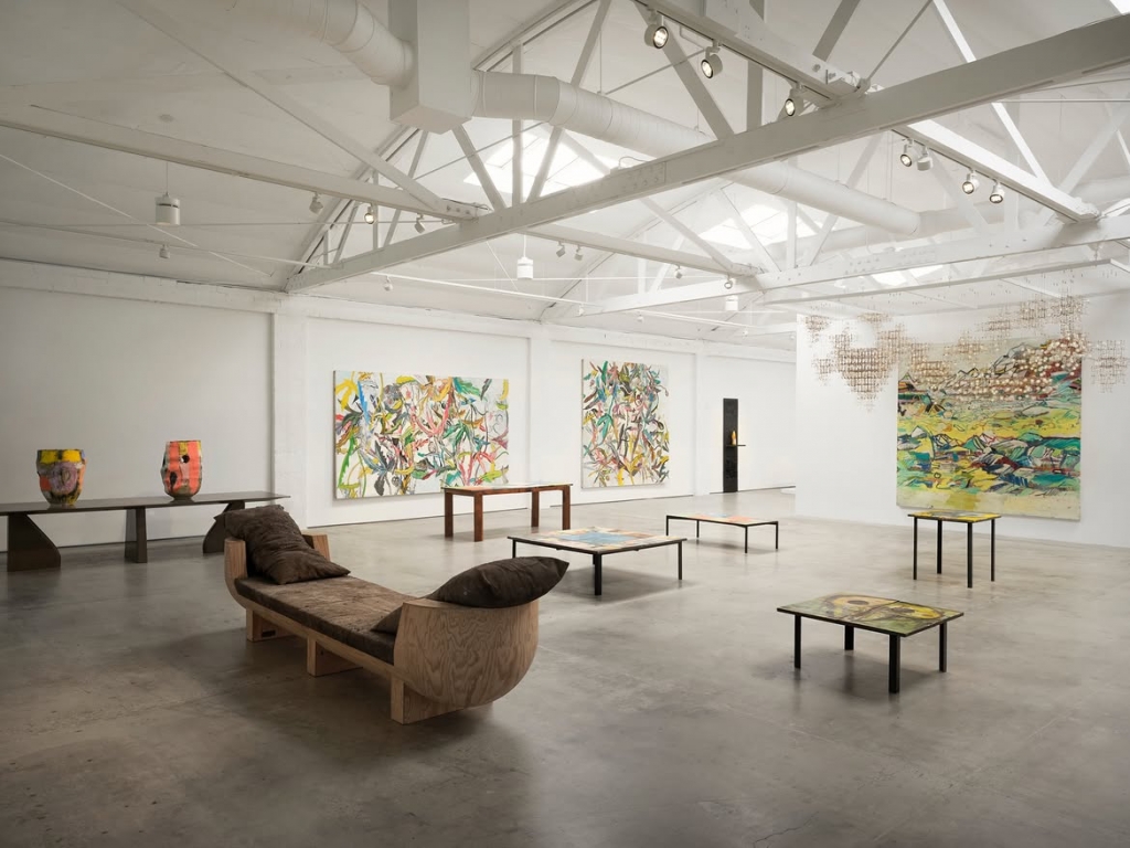 A spacious, white industrial art gallery room displaying abstract colorful paintings, modern wooden benches, and ceramic vases at PAD Saint-Tropez 2026.