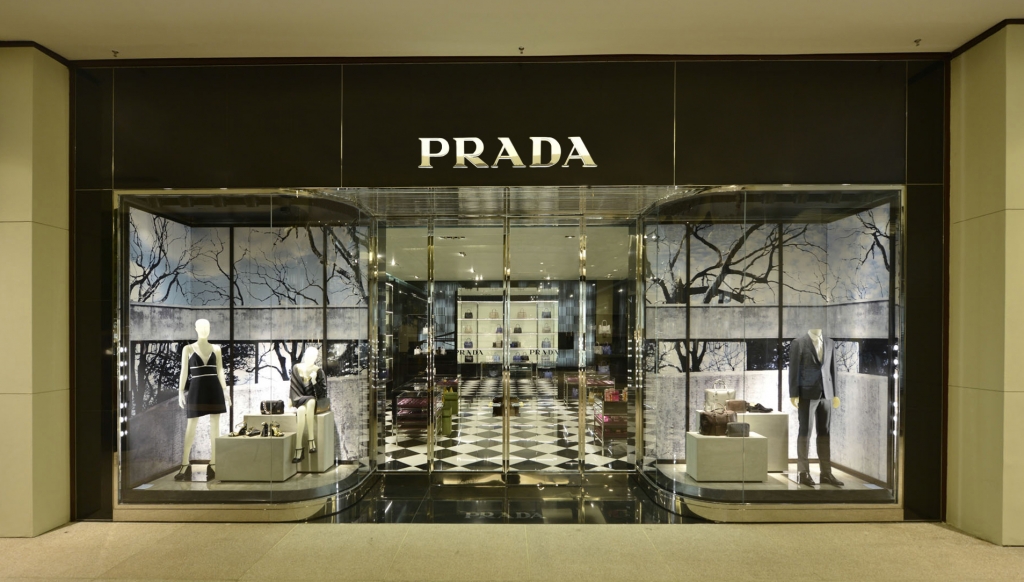 Vitrine da loja Prada com manequins vestidos, piso em xadrez preto e branco e cenário decorativo de árvores ao fundo - lojas de luxo em São Paulo