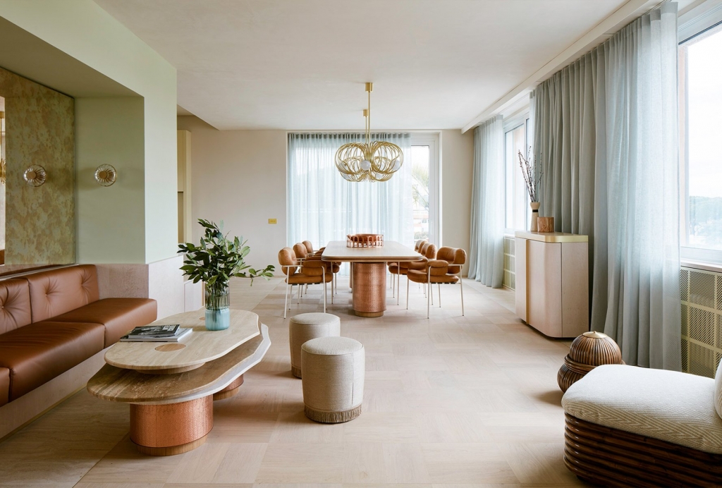 Soft pastel dining and lounge area with a terracotta leather sofa, sculptural travertine coffee table, rattan-based dining table, caramel leather chairs, and a gold ring chandelier. interior designers