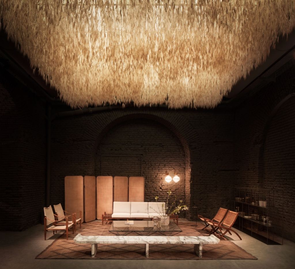 Moody exhibition space with exposed brick arches, a fringed straw/rope ceiling installation, leather campaign chairs, a marble bench, and a cream sofa. interior designers
