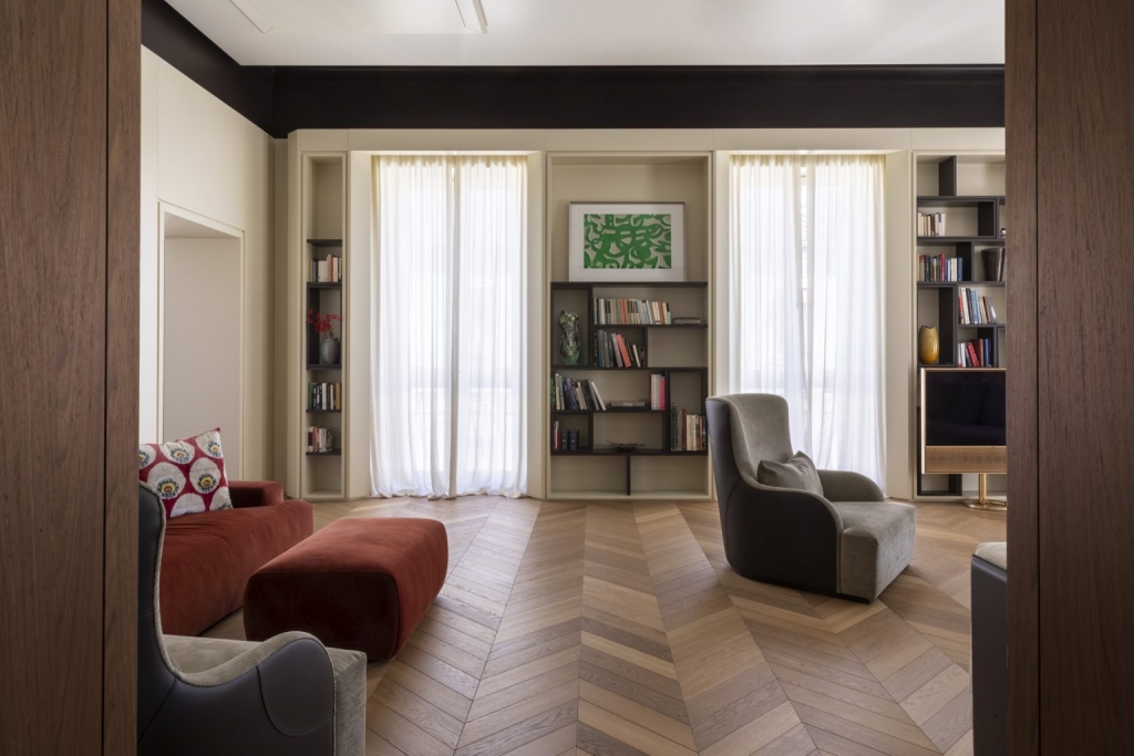 Elegant living room with a red velvet sofa and ottoman, dark leather wingback chairs, herringbone parquet flooring, and built-in bookshelves flanking sheer-curtained French doors. interior designers