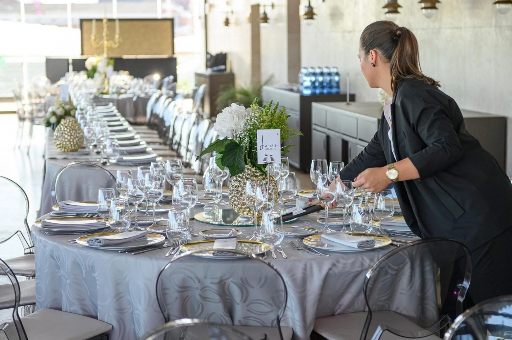 Event coordinator arranging place settings and wine glasses on an elegantly set banquet table with gray linens, gold-rimmed plates, and floral centerpieces, with a long row of additional set tables in the background. private sunset