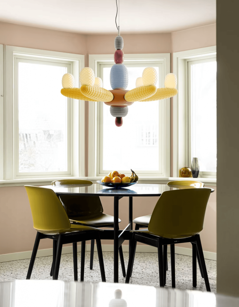 Playful pink dining nook with a sculptural multicolor pendant light, round black table set with mustard-yellow chairs, and terrazzo flooring by a bay window. interior designers
