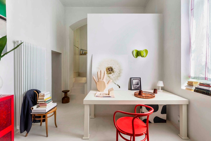 Playful white study nook with a red bentwood chair, whimsical sculptural objects, a dandelion photograph, and a glimpse of a staircase through an archway. interior designers