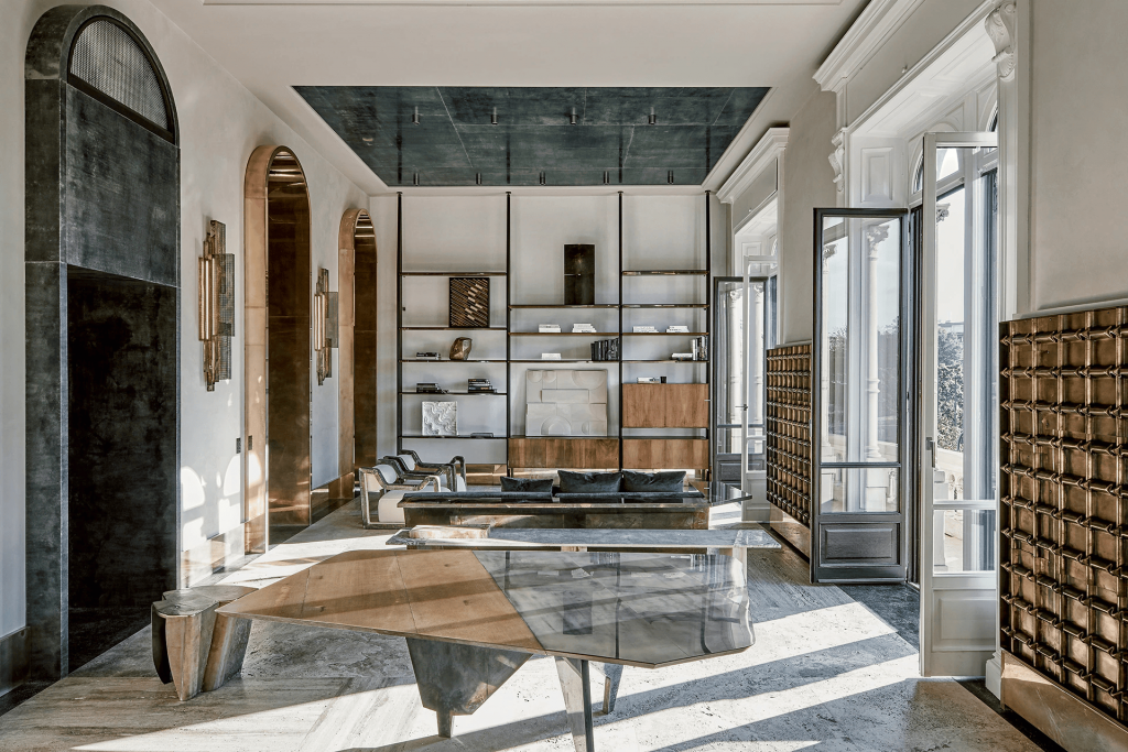 Refined Roman apartment salon with arched bronze-mirrored niches, a slate-blue ceiling panel, open shelving displaying art objects, and a sculptural bronze coffee table. interior designers
