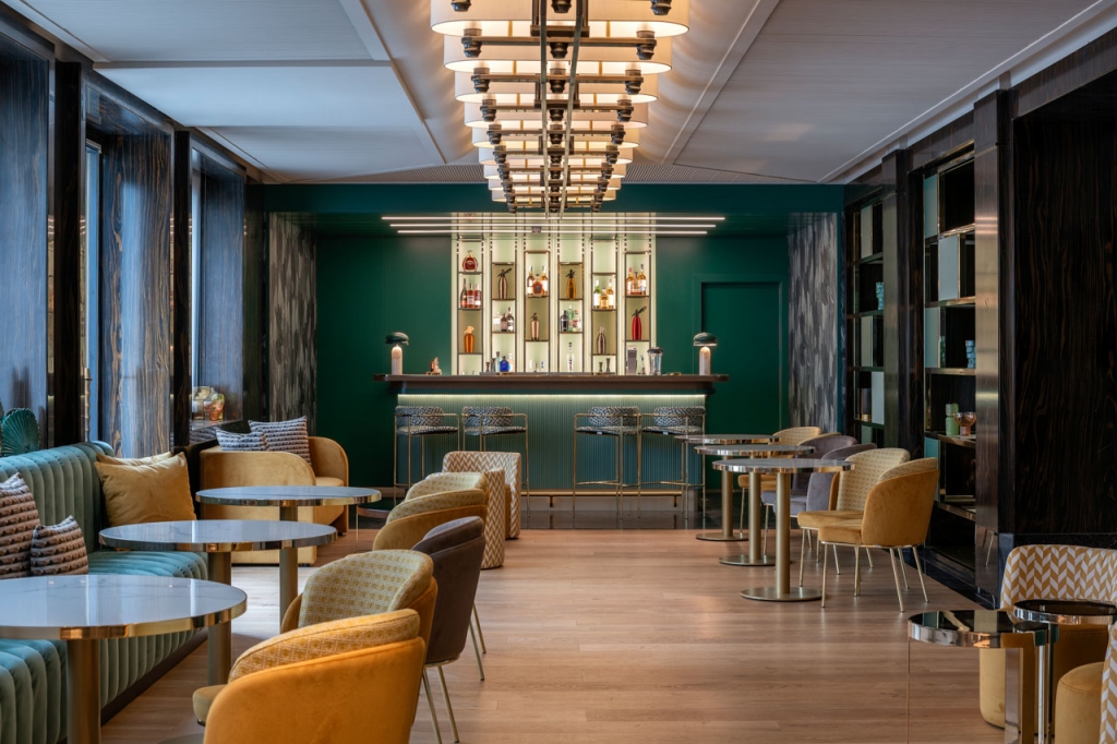 Hotel bar lounge with emerald green walls, gold-trimmed marble side tables, mustard velvet armchairs, a teal channel-tufted banquette, and a tiered drum chandelier above a backlit liquor display. interior designers