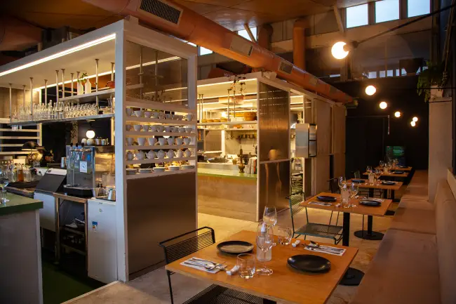 Cozinha aberta com prateleiras de louças e copos, balcão de aço inox e mesas simples de madeira com pratos pretos ao fundo - melhores restaurantes em São Paulo