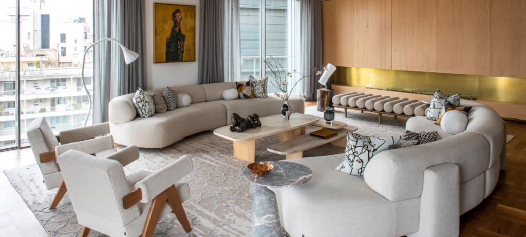 Bright contemporary living room with a curved boucle sectional sofa, patterned throw pillows, a stone coffee table, wood-paneled wall with brass strip, and a framed portrait painting. interior designers