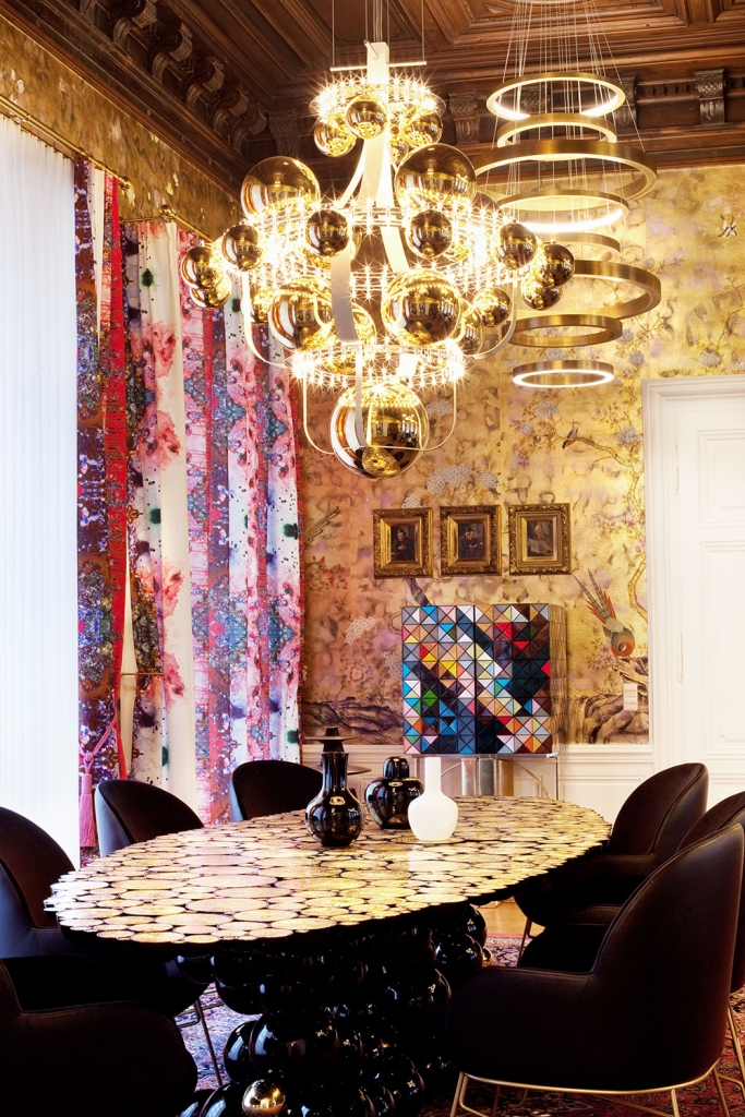 Opulent dining room under a carved wood ceiling with a golden sphere chandelier and a ring-style pendant light, a mosaic oval dining table, black leather chairs, gilded floral wallpaper, framed portraits, and a colorful geometric cabinet, denis kosutic