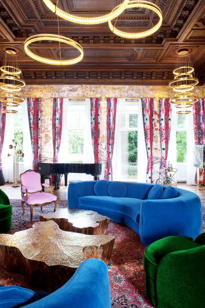 Ornate palace living room with carved wood coffered ceiling, gold ring chandeliers, floral curtains, a grand piano, a royal blue curved velvet sofa, emerald green armchairs, and sculptural bronze coffee tables on a Persian rug, denis kosutic