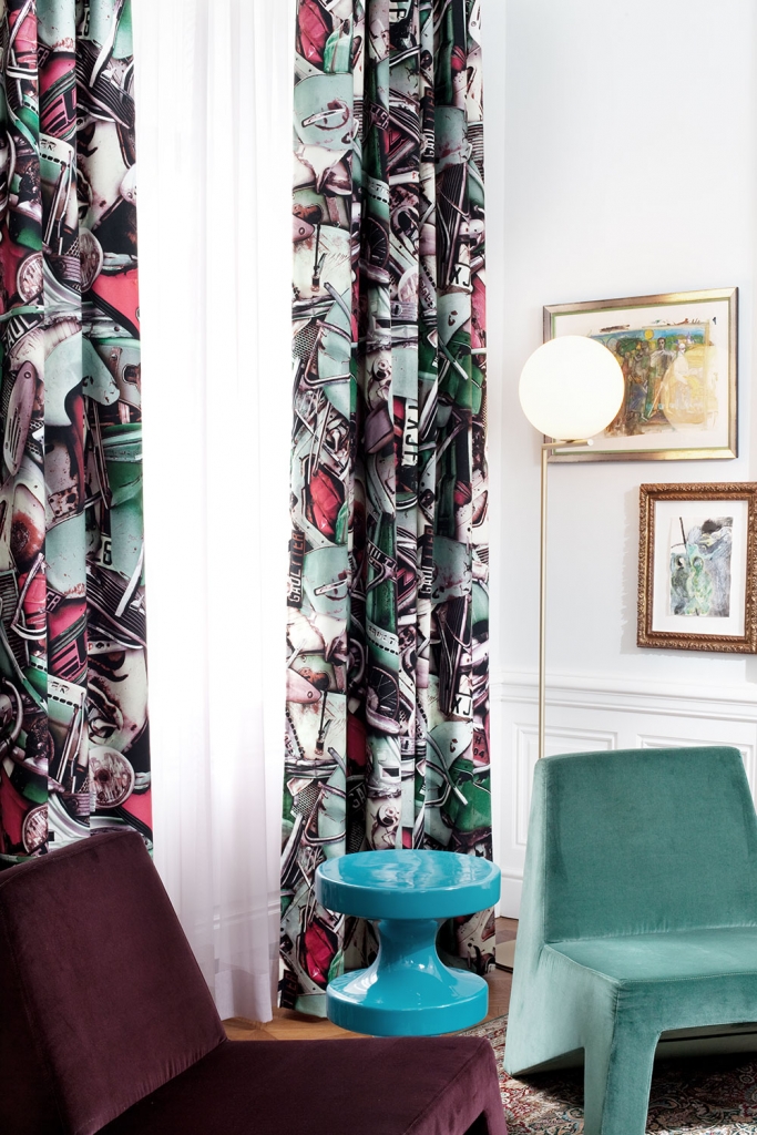 Detail of graphic printed curtains with car and motorcycle motifs beside a teal velvet chair, a maroon velvet chair, a turquoise pedestal side table, a round glass floor lamp, and framed abstract artworks on a white paneled wall, denis kosutic