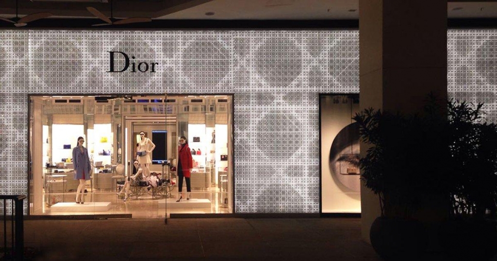 Fachada noturna da loja Dior com padrão decorativo iluminado na parede e vitrine mostrando manequins com roupas coloridas - lojas de luxo em São Paulo