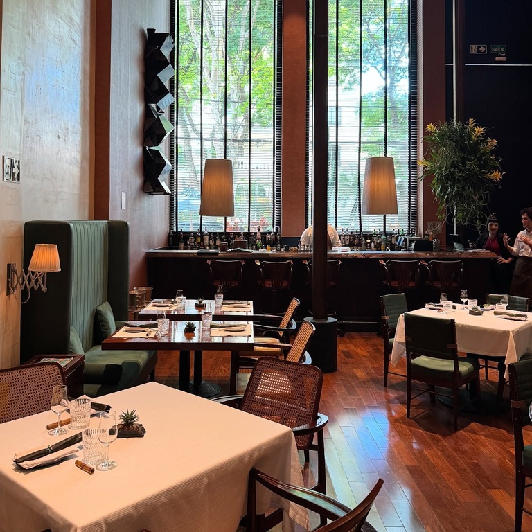 Salão de restaurante com mesas cobertas por toalhas brancas, cadeiras de palhinha, banco verde estofado e balcão de bar ao fundo com garrafas iluminadas - melhores restaurantes em São Paulo