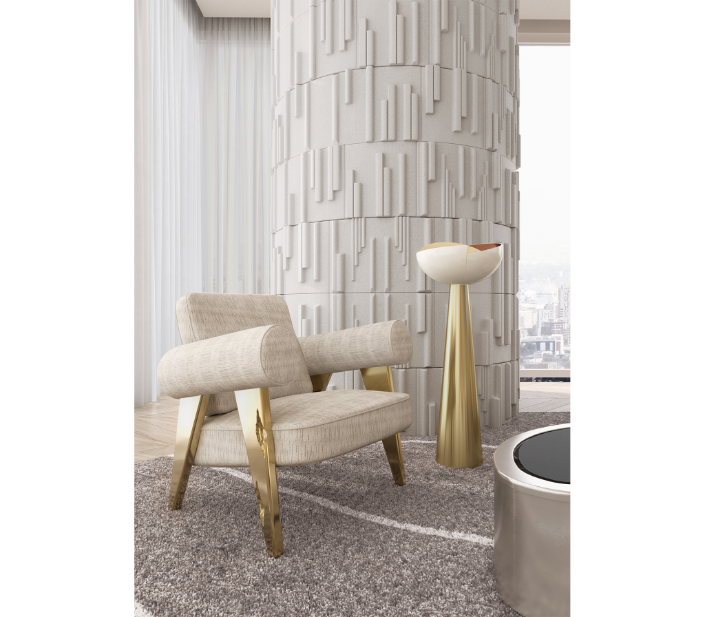 Cream textured accent chair with bolster-style arms on angular gold legs, beside a gold conical table lamp and a textured white column, with a city view in the background- luxury armchair.