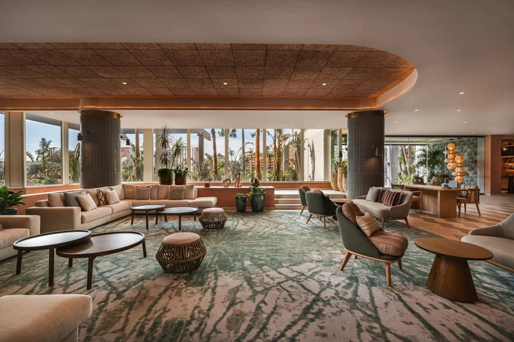 Resort-style hotel lobby lounge with a curved textured ceiling, terrazzo-look green rug, rattan furniture, and floor-to-ceiling windows overlooking palm trees. interior designers