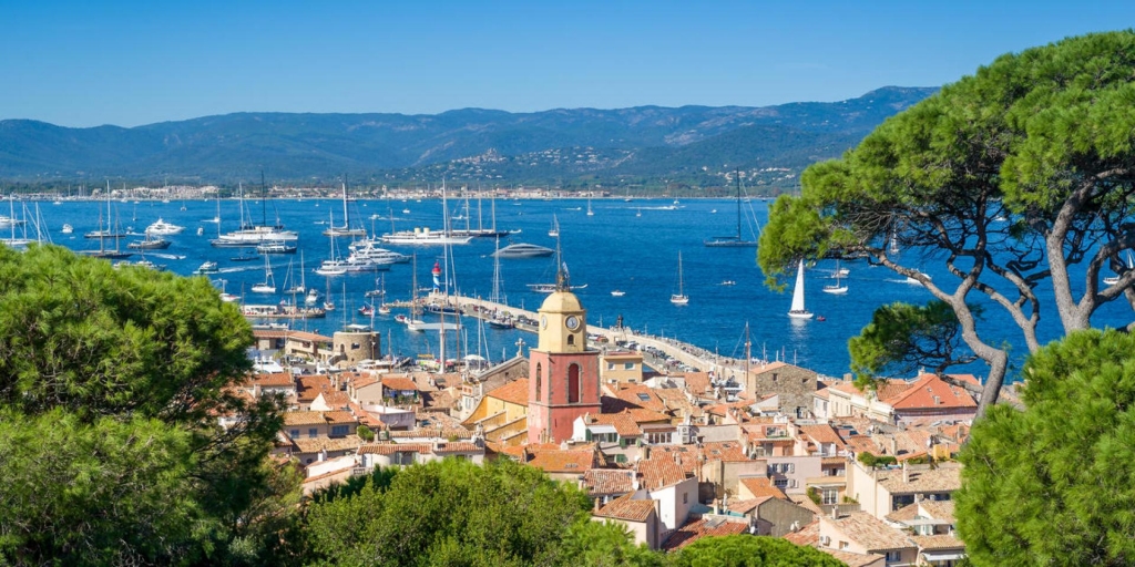 High-angle panoramic view of a coastal town harbor filled with yachts and sailboats next to a red-and-yellow clock tower at PAD Saint-Tropez 2026.