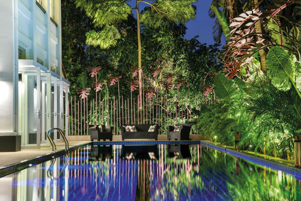 Nikki Hunt Interior Design, Exterior of the house with a swimming pool and a seating area featuring black sofas and armchairs with colourful cushions, surrounded by lush vegetation in a tropical style