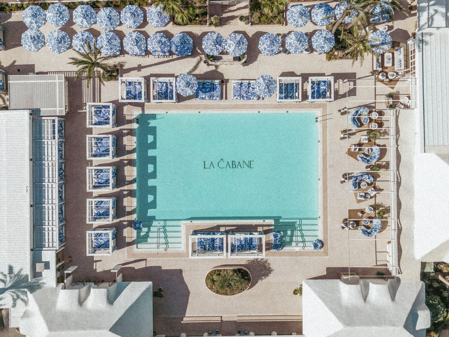 Relaxed luxury at La Cabane by Dolce&Gabbana featuring a large swimming pool surrounded by blue-and-white sun loungers, striped parasols, palm trees, and the Mediterranean Sea in Marbella.
