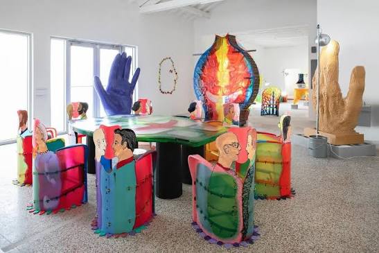 Whimsical art gallery installation with a round table surrounded by illustrated cutout figure chairs in bright colors, alongside oversized sculptural hands and a flame-shaped stained-glass piece. INTERIOR DESIGNERS