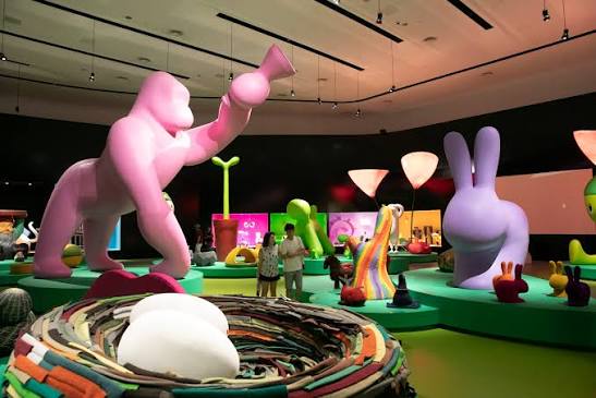 Colorful immersive art installation featuring giant sculptural figures (a pink gorilla, purple rabbit) on green display platforms, with a woven multicolor nest sculpture in the foreground. interior designers