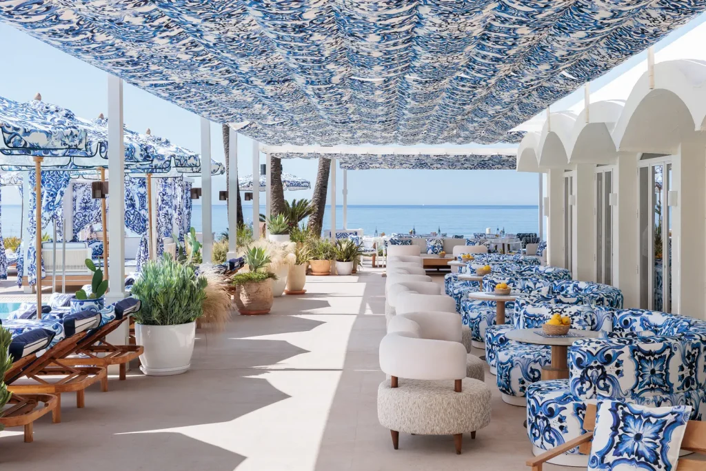 Relaxed luxury at La Cabane's open-air restaurant with blue-and-white table settings, woven chairs, Mediterranean plants, and sea views designed by Dolce&Gabbana.