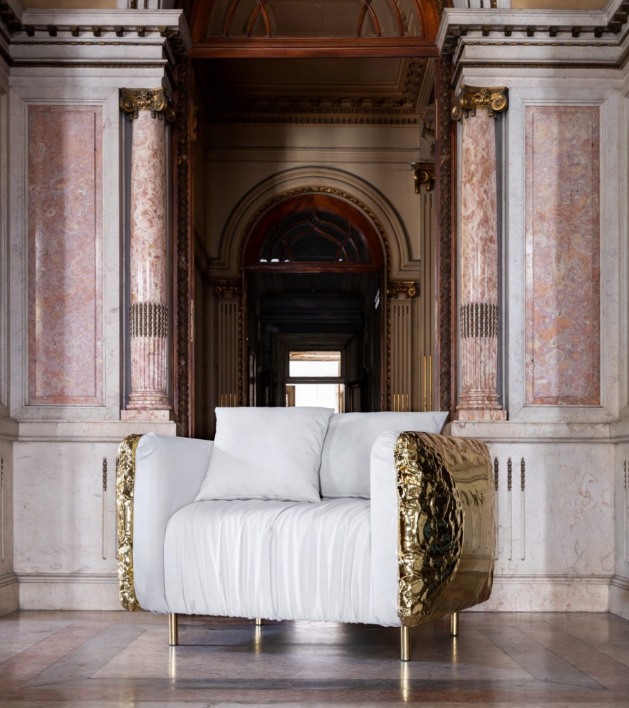 Beige Imperfectio armchair with polished brass side panels displayed in a grand classical interior with marble columns and ornate architectural details, luxury armchair.