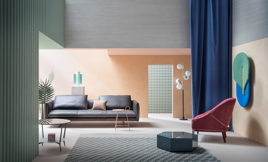 Playful, color-blocked showroom set with a grey leather sofa, coral velvet chair, geometric wall art, navy curtain, and a checkered rug on a sage-and-peach backdrop. interior designers