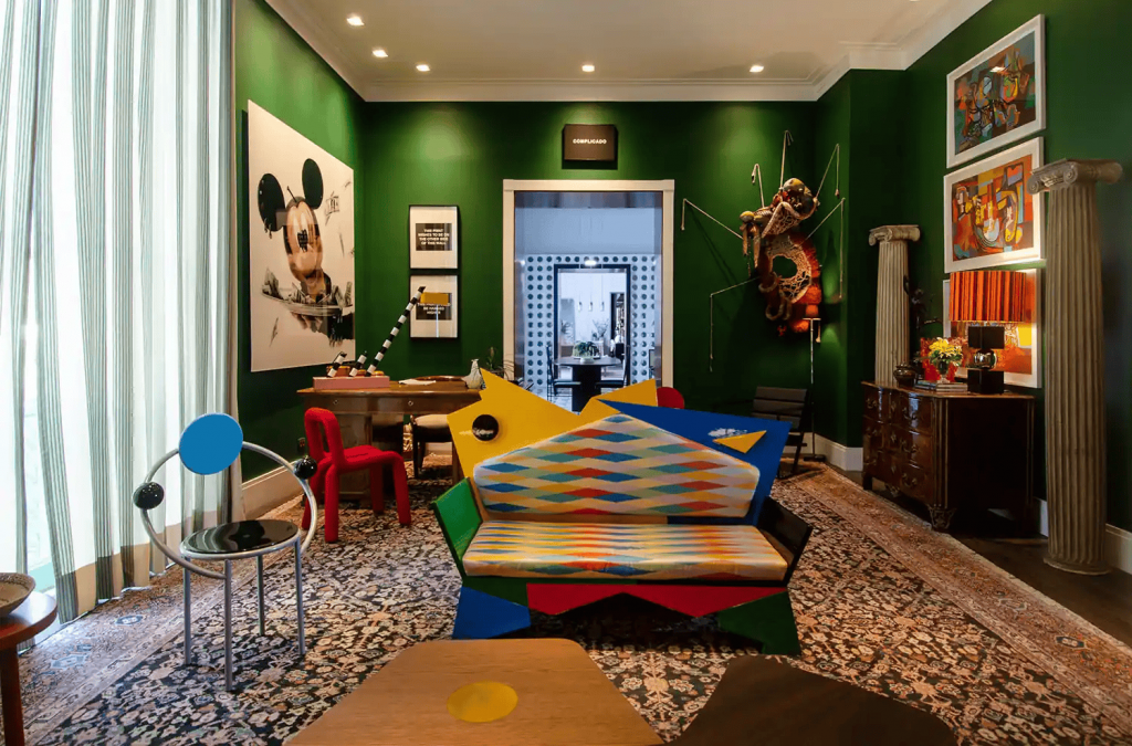 Eclectic emerald-green living room with Memphis-style geometric sofa in primary colors, postmodern chrome and glass side chairs, gallery wall of pop art, and an antique wooden dresser. interior designers