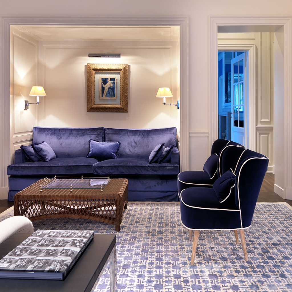 Classic cream living room nook with a navy blue velvet sofa and matching piped armchair, a woven rattan coffee table, gilt-framed artwork, and patterned area rug. interior designers