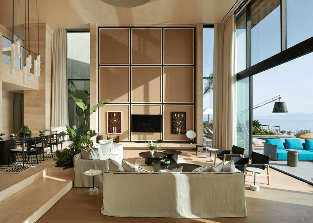 Double-height luxury villa living room with a large coffered stone accent wall, tribal-inspired artwork, linen slipcovered sofas, and sliding glass doors opening to a sea-view terrace with turquoise loungers. interior designers