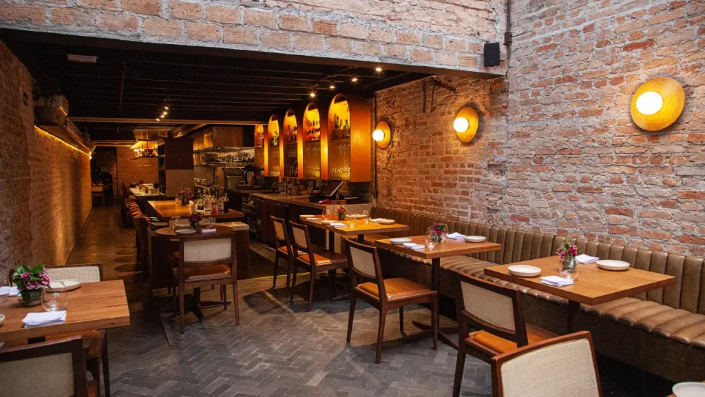 Restaurante com paredes de tijolo à vista, nichos iluminados em tom âmbar atrás do bar, mesas de madeira e bancos estofados em couro - melhores restaurantes em São Paulo