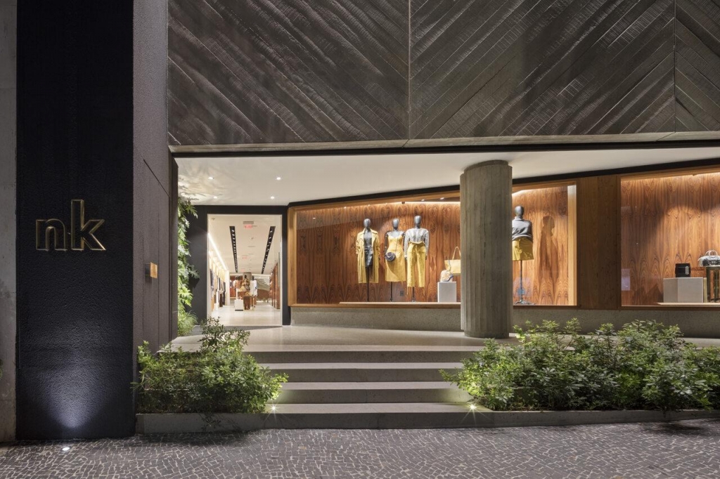 Fachada noturna do shopping NK com revestimento de madeira em espinha de peixe, escadaria de acesso e vitrine com manequins vestidos - lojas de luxo em São Paulo