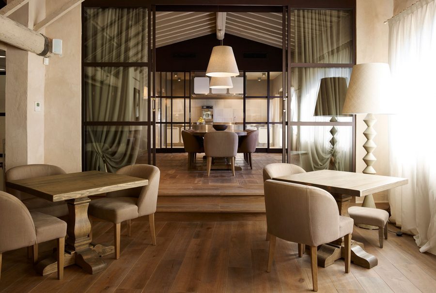Rustic-chic Tuscan restaurant interior with exposed wood beams, linen café chairs, wrought-iron framed glass partitions, and diamond-based table lamps. interior designers