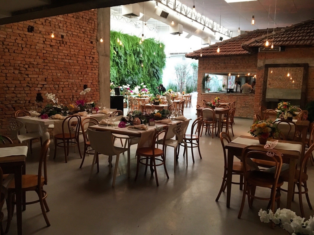 Salão amplo com parede de tijolo aparente, jardim vertical, mesas postas para evento com flores coloridas e cadeiras de madeira estilo bistrô - melhores restaurantes em São Paulo
