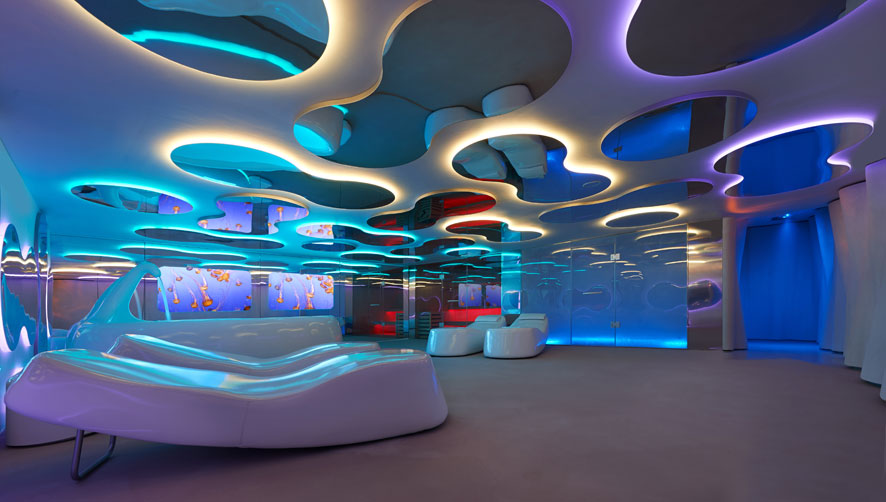 Futuristic spa lounge with organic cloud-shaped ceiling cutouts, multicolor LED mood lighting, mirrored walls, and sculptural white lounge seating. interior designers